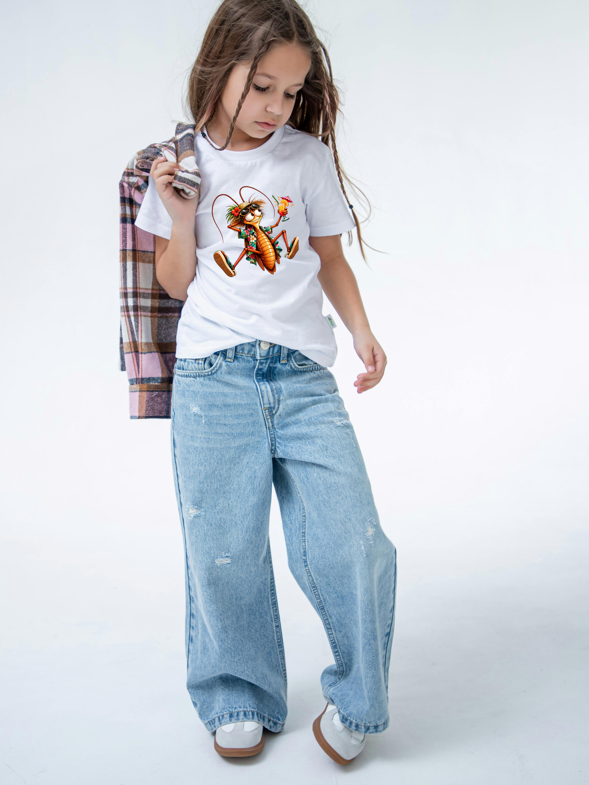 design mock ups. Design can be used on t-shirt, mug, wall paper, sticker, and more. A young girl modeling a white graphic t-shirt with a cartoon monkey, paired with wide-leg light blue jeans and holding a plaid shirt under her arm, set against a plain white studio background.